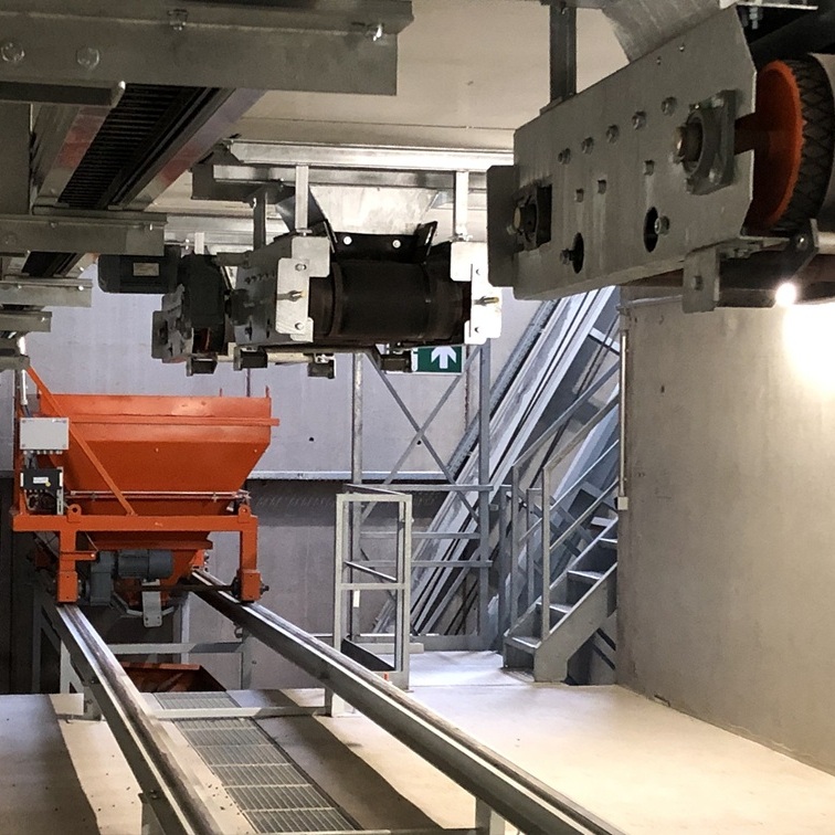 Orangener Waagen auf Schienen in Betonbau. Darüber hängend verzinkte Förderbänder.