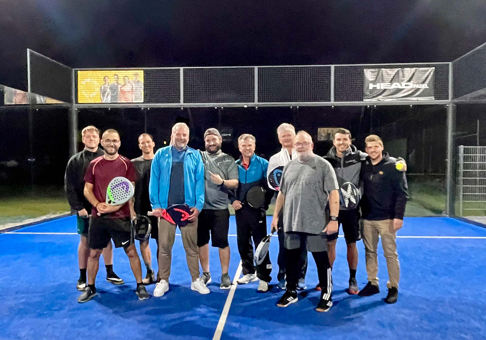 Gruppe auf blauem Padelplatz bei Nacht, mehrere Personen halten Schläger und stehen vor Glaswänden.