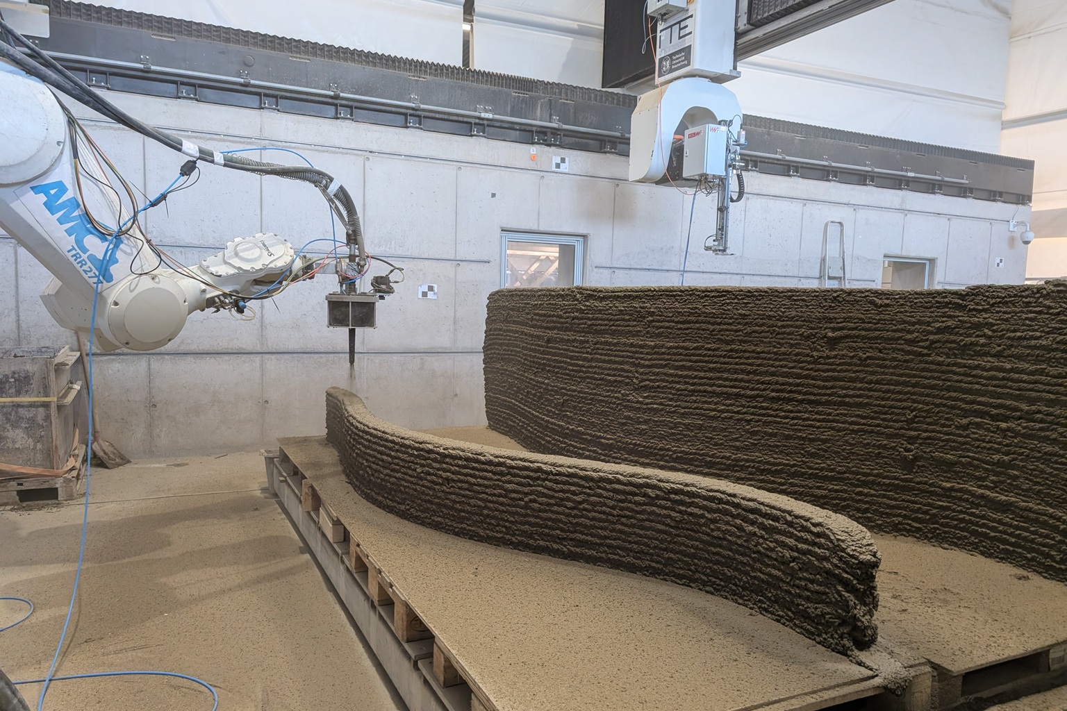 Un robot industriel imprime du béton couche par couche pour former un mur courbé dans un atelier; l'impression 3D du béton est mise en œuvre.
