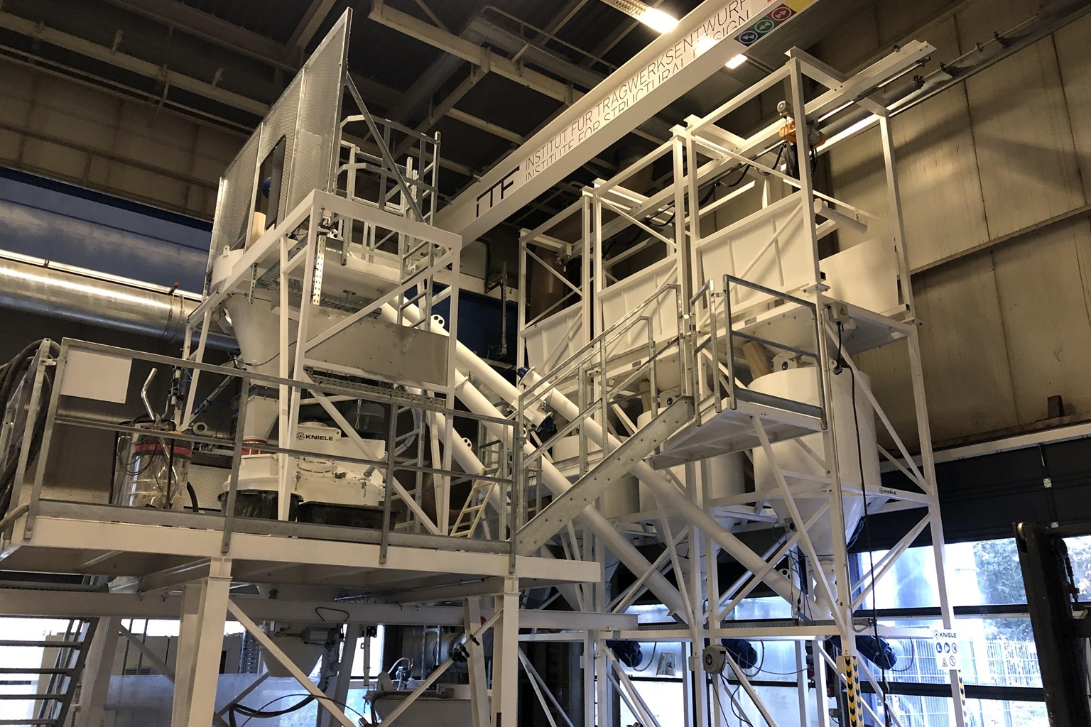 Grande installation métallique de mélange et de transport, avec des escaliers et des plates-formes, dans un hall industriel; installation d'essais techniques visible.
