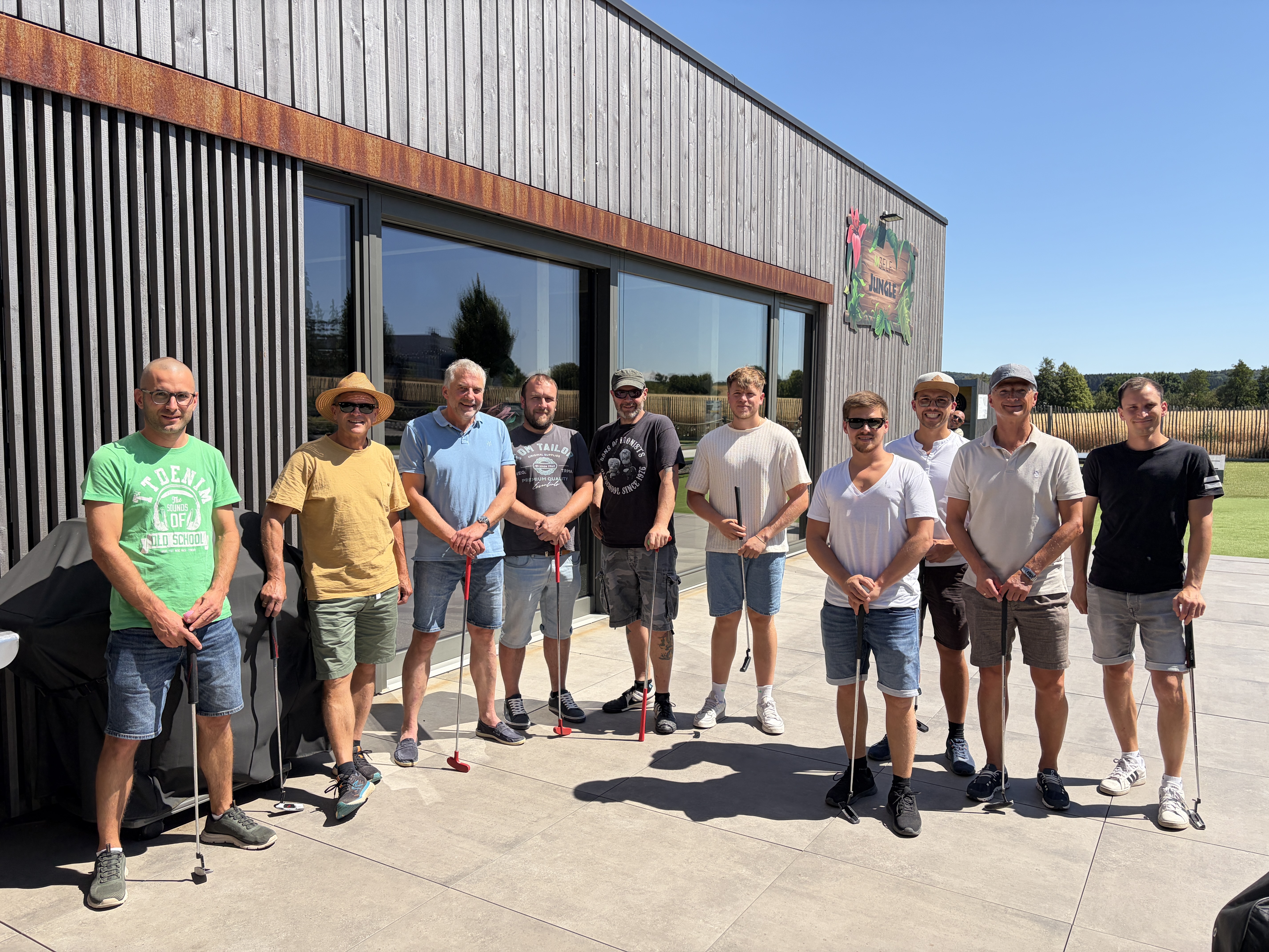 Un groupe de dix personnes munies de clubs de minigolf devant un bâtiment moderne en bois, sous un soleil radieux.