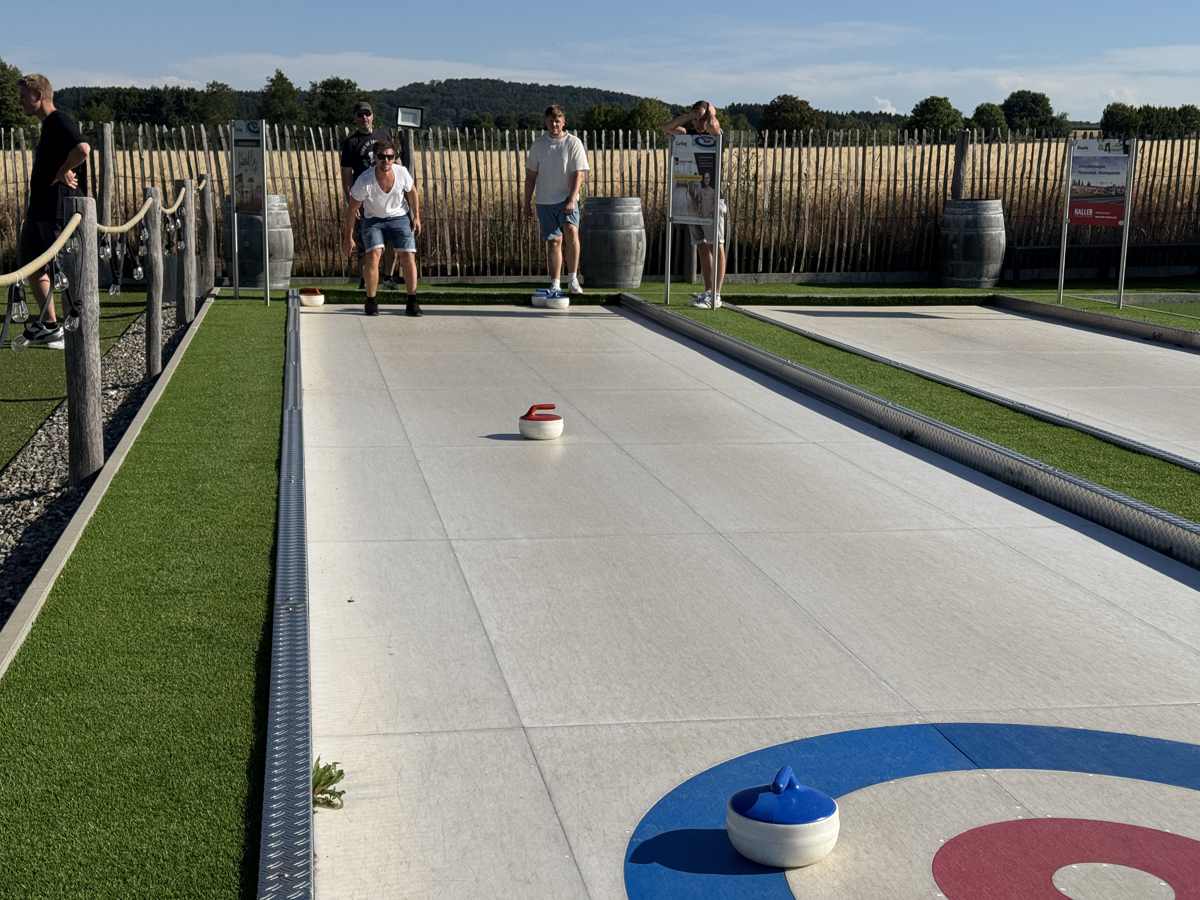 Des personnes jouent au curling sur une piste en plein air, au milieu des champs et des arbres, par temps ensoleillé.