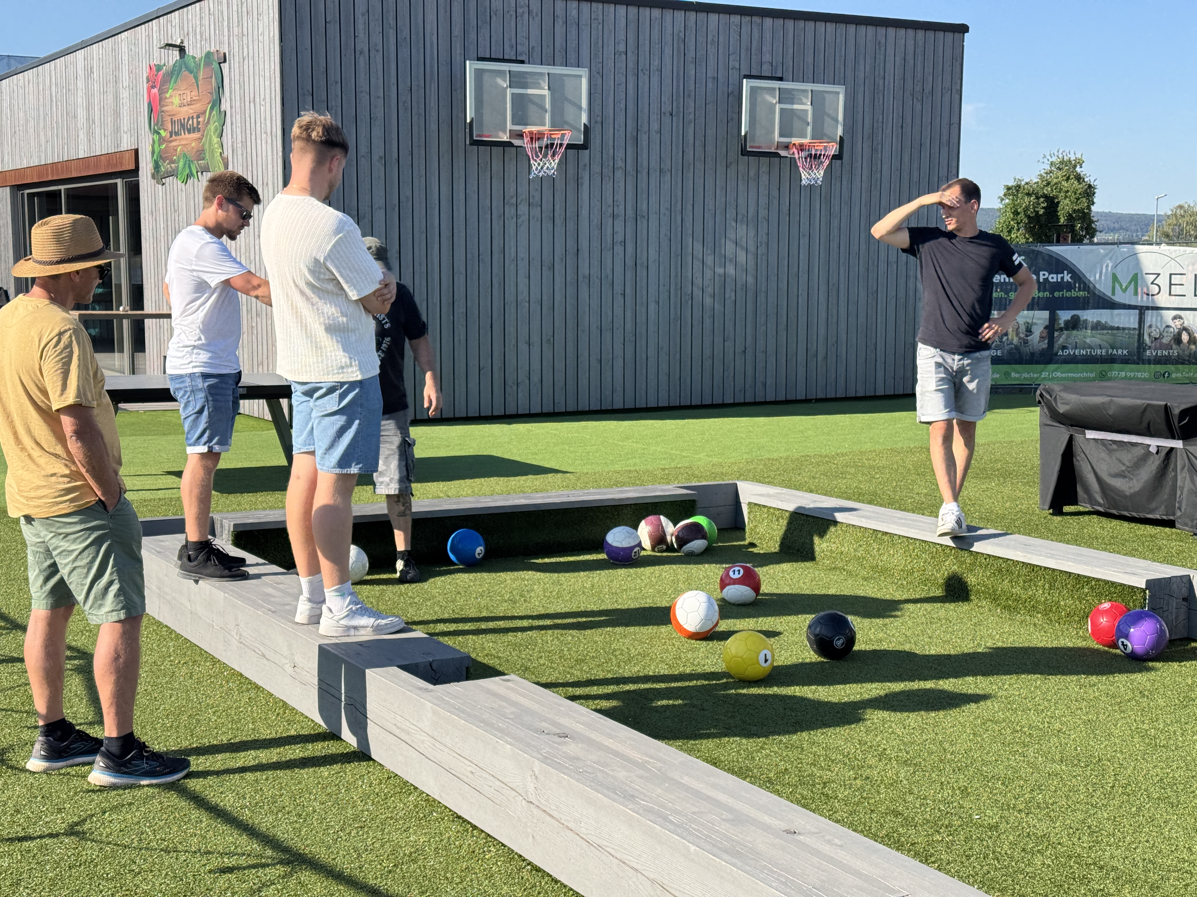 Plusieurs personnes jouent au billard-foot sur un terrain en plein air, avec de grosses balles et des bandes en bois.