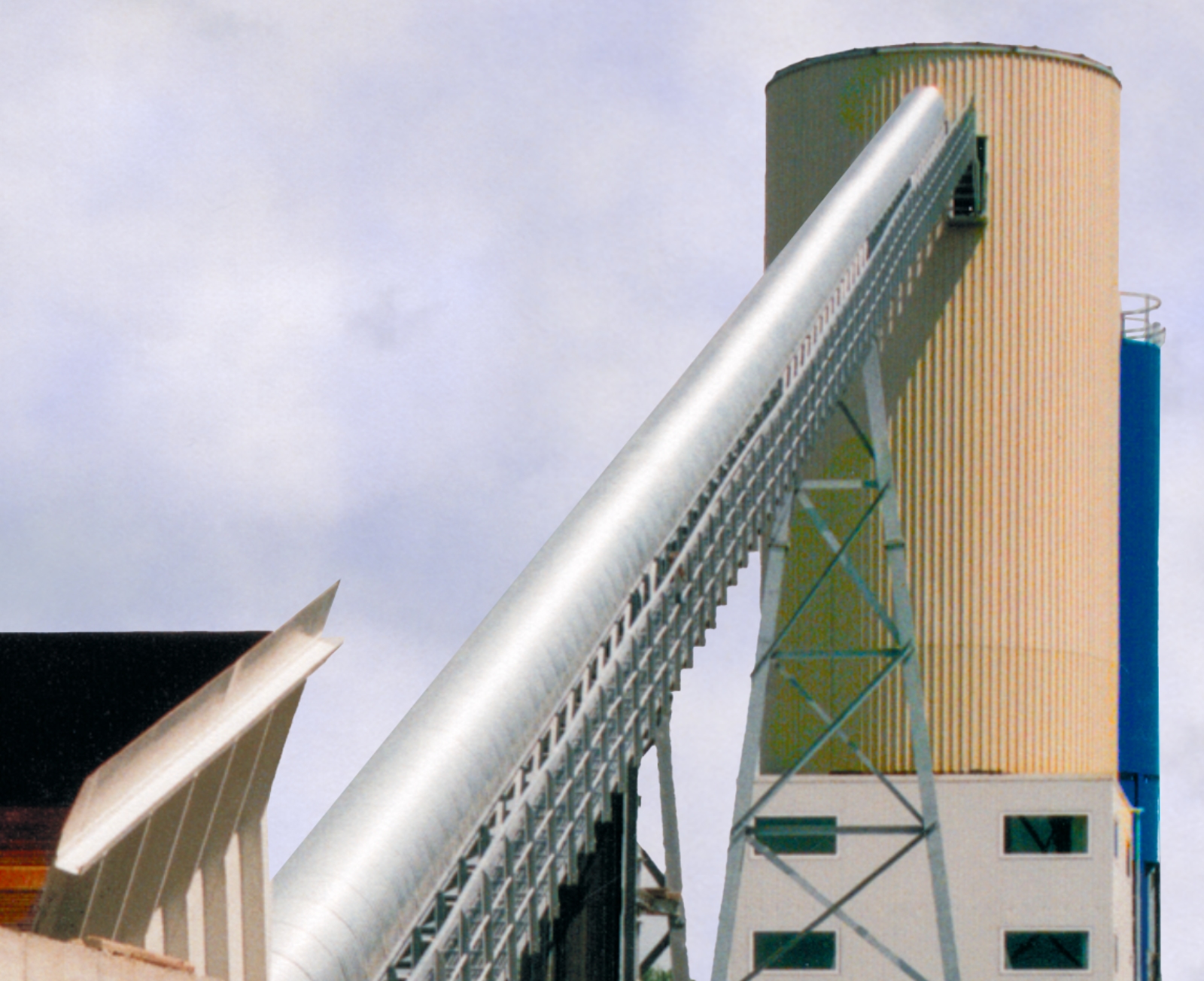 Long inclined conveyor belt with a semicircular cover from a feed hopper to a high cylindrical mixing tower.
